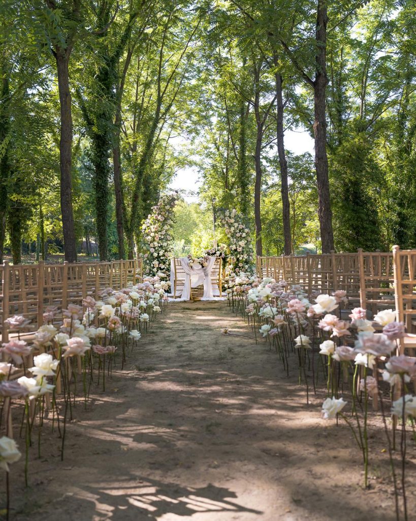 Cerimonia all'aperto con sedie chiavari dorate e arco floreale bianco tra gli alberi
